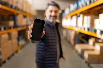 Warehouse worker holding smartphone blank screen mockup. Logistics manager using app for inventory tracking. Digital transformation in industry and supply chain background.
