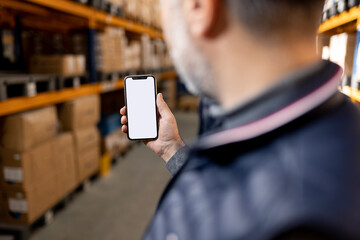 Warehouse worker holding smartphone blank screen mockup. Logistics manager using app for inventory tracking. Digital transformation in industry and supply chain background.