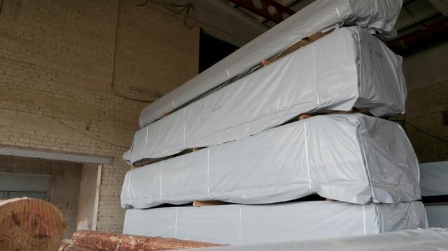 Large wrapped timber beams and unprocessed logs are stored in an industrial warehouse with brick walls and exposed beams. Dim lighting, no visible logos or faces