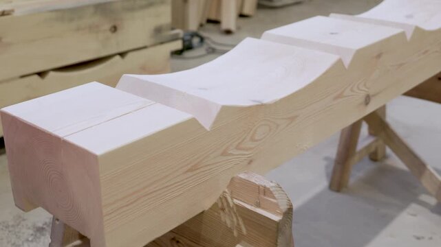 Two workers in safety gear position a carved wooden beam on wooden sawhorses inside a carpentry workshop. No visible logos or faces