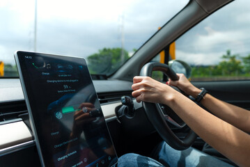 Asian woman driver electric car along a scenic winding road, surrounded by lush greenery. The digital touchscreen displays the electric car battery level and charging options. Future energy concept.