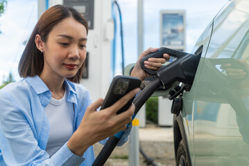 Asian woman driving electric car looking at time on smartwatch while waiting for electric car to charge for safe next drive. Future energy concept.
