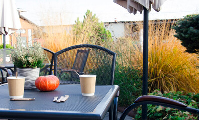 Cozy outdoor cafe table set in an autumn garden with two takeaway coffee cups, a small pumpkin, and potted heather plant.