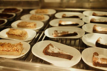 Assorted cake slices arranged on white plates inside a bakery display case. Sweet dessert selection showcasing variety, freshness and cafe pastry menu.