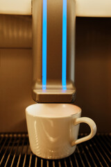 White ceramic cup placed under a modern coffee machine before brewing. Blue indicator lights glow, creating a clean minimalist mood and professional coffee preparation concept.