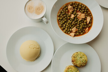 Close view of green pea stew with chicken pieces served on white plates with mashed potatoes and vegetable patties. Warm homemade lunch concept, casual dining atmosphere.