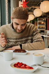 A man sits at a cafe table with cappuccino and desserts, holding a fork and looking down in a warm modern cafe interior.