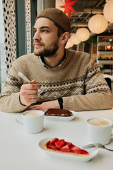 A man sits at a cafe table with cappuccino and desserts, holding a fork and looking away from the window in a cozy modern interior.