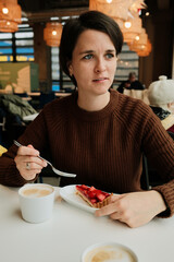 A woman sits at a cafe table with cappuccino and a strawberry tart, holding a fork and looking to the side in a warm interior.
