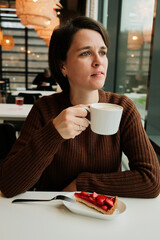 A woman holds a cup of cappuccino near a window with a strawberry tart on the table in a modern cafe interior.