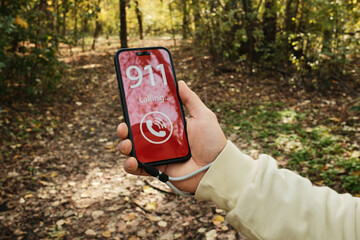 Hand holding a smartphone showing a 911 emergency call screen in a forest path. Concept of danger, urgent help request, outdoor accident risk and emergency communication in nature.