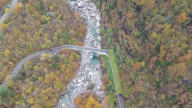 Aerial view of a bridge over a river flowing through Lavertezzo surrounded by autumn trees with roads on either side, Verzasca, Ticino, Switzerland.