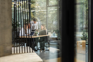 A view through a glass partition shows a modern, busy office interior. Business people are working at desk