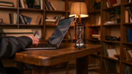 Person typing on laptop next to hourglass in a library setting with bookshelves © shine.graphics