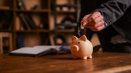 Person in suit putting coin into a piggy bank on a wooden desk with an open book