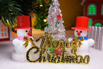 Two white snowmen with letters and a Christmas tree against the background of Christmas houses
