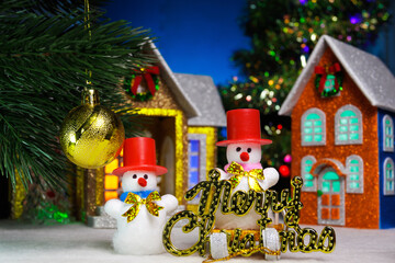 A snowman and a snowman on a sled with letters next to a ball on a Christmas tree branch against the background of houses