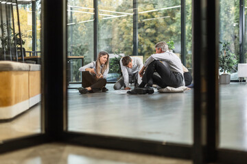 Happy successful business team sitting on floor in office, having fun at work, diverse coworkers chatting, laughing at colleagues joke, positive employees enjoying break at work together