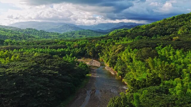 Aerial view of a winding river cutting through lush green forests and rolling hills, under a dramatic cloudy sky, Maasin, Western Visayas, Philippines.