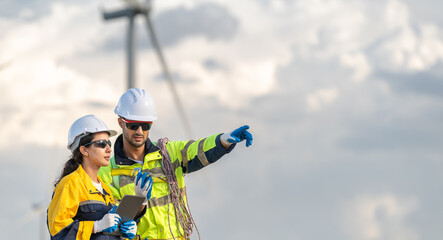 Two renewable energy engineers in safety gear discussing data on laptop at wind turbine field. Male and female inspection engineer team working at windmill site for sustainability project.