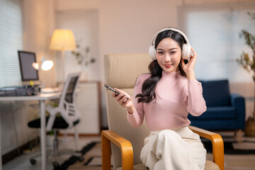 Young asian woman listening to music on headphones and holding smartphone in home office