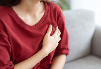 Young woman in red shirt clutching her chest while suffering from acute pain or heart attack symptoms at home, health emergency and cardiovascular disease concept.