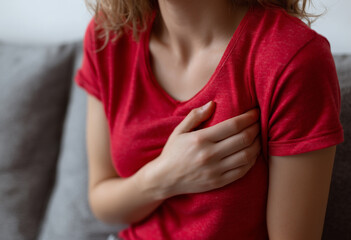 Young woman clutching her chest in pain while sitting on a sofa at home, concept of heart attack symptoms, anxiety, and cardiovascular health issues for medical awareness