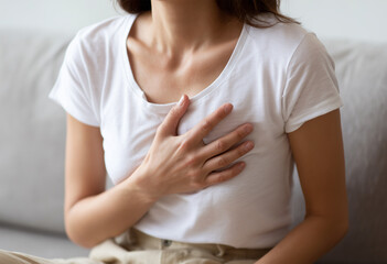 Woman clutching her chest in pain while sitting on a couch indoors, representing symptoms of heart attack, anxiety, or respiratory distress for health and medical topics