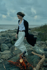 Woman walks along a rocky shoreline by the sea in windy weather, wearing white wide-leg pants, a...
