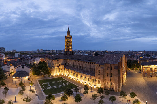 Aerial view of Saint Sernin Basilica bathed in the soft glow of twilight, its brick facade a warm contrast to the deepening blue of the evening sky, Toulouse, Occitanie, France.