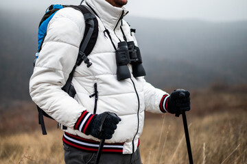 Close up photo of hiker in warm clothing with backpack and binoculars holding hiking poles while...