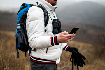 Close up photo of hiker in warm clothing using smartphone while hiking in nature with backpack, hiking poles and binoculars on foggy winter day.