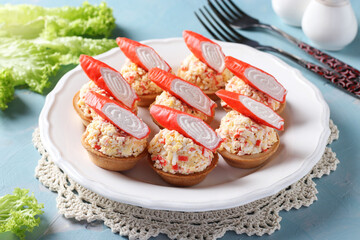 Tartlets with crab sticks, eggs, cheese and cucumber on white plate on blue background