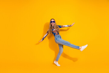 Young woman dancing joyfully in a bright yellow studio wearing jeans sneakers and sunglass casual fashion vibe