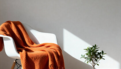 White modern chair with orange knitted blanket and green plant. Minimalist home interior background with sunlight and shadow. Copy space for text