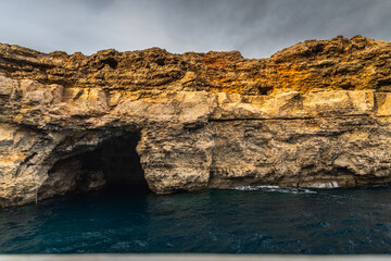 Dramatic coastal view of the Santa Maria Caves in Comino, Malta, featuring rugged limestone cliffs and sea caverns overlooking the deep blue Mediterranean Sea. Travel to Malta.