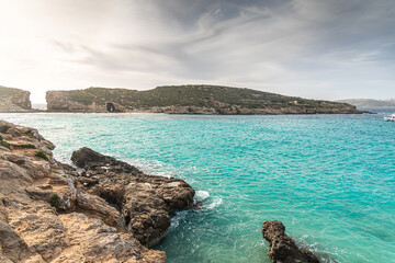 Malta Blue Lagoon landscape. Stunning view of the Blue Lagoon in Comino, Malta, showing the famous turquoise crystal clear waters and the rocky islet of Cominotto under a cloudy sky.
