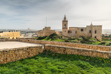 Gozo citadella. Scenic view of the ancient stone fortifications and rooftops within the Cittadella of Victoria, Gozo, showing green grass and traditional limestone architecture.