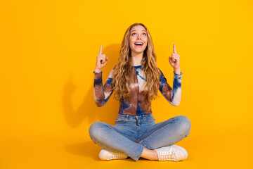 Young girl with blonde hair sits cross legged on a bright yellow background pointing up with both hands smiling and stylish casual fashion