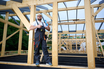 Construction worker taking a break and drinking water at wooden house frame construction site. Builder resting with hard hat and water bottle.