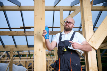 Construction worker taking a break and drinking water at wooden house frame construction site. Builder resting with hard hat and water bottle.