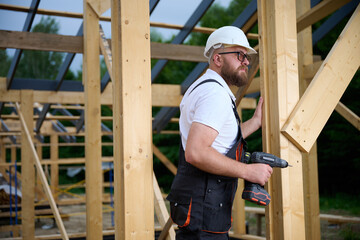 Construction worker using an electric screwdriver while building a wooden frame structure. Man in protective helmet, gloves. Concept of construction, carpentry, engineering and building process.