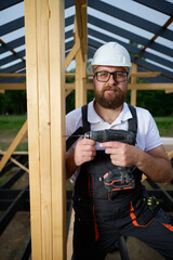 Construction worker using an electric screwdriver while building a wooden frame structure. Man in protective helmet, gloves. Concept of construction, carpentry, engineering and building process.