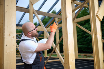 Construction worker checking wooden beam alignment at timber frame house site. Builder in safety gear holding helmet and using tools while working on wooden structure outdoors.