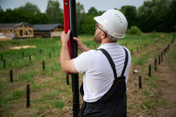 Construction worker installing screw pile foundation at building site using water level. Professional builder in helmet aligning metal ground screw for house base installation.