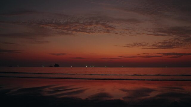 Stunning sunset over the beach in Ranong, Thailand, featuring a deep orange sky, calm waves reflecting on the sand, and distant fishing boat lights on the horizon.