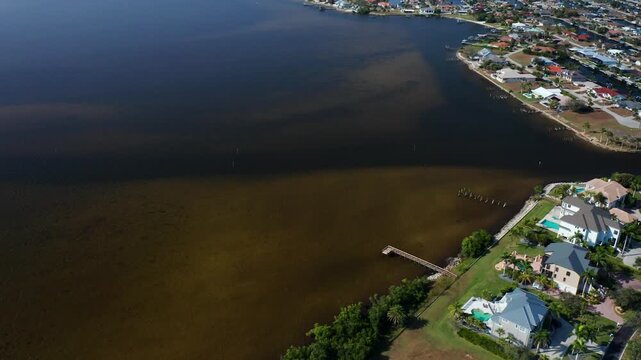 Waterfront houses with private docks overlook a calm brownish coastal bay, surrounded by green patches and a sprawling residential neighborhood with clear skies.