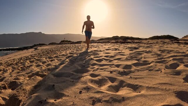 LOW ANGLE VIEW, LENS FLARE, SLOW MOTION: Clumsy man trips over while running and falls onto sandy beach. Sand flies in all directions in golden light while man lies on ground after spectacular fall.