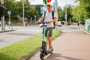 Fototapeta premium Man moves on an electric scooter down a city street with traffic and pedestrians. It is a sunny day and he carries a backpack