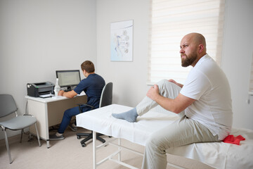 Male patient being examined in clinic for knee pain. Male suffering from knee pain osteoarthritis. Close up man holding his knee due to physical injury while sitting in room.